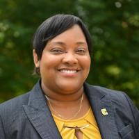 Rosite Delgado Black woman with dark grey blazer and yellow top smiling in front of a blurred green background
