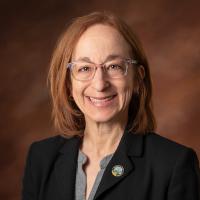 Jenni Berz While woman in black blazer with grey top and glasses in front of a brown background