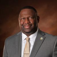 Dennis Clark Black male in grey suit and gold tie in front of a brown background