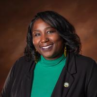 Raquetta Dotley Black woman with black blazer and green top in front of a brown background