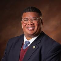 Ron Elliot Black male with blue blazer, red sweater, and light blue tie with glasses in front of a brown background