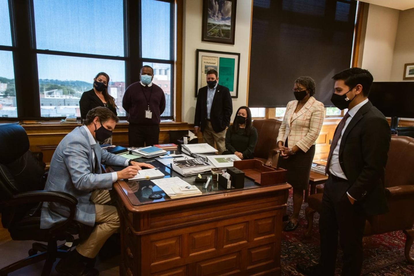 En City Hall, el Alcalde Tim Kelly y su equipo, vestidos con trajes y mascarillas, se reúnen alrededor de un escritorio para firmar una legislación importante para los presupuestos de Chattanooga.