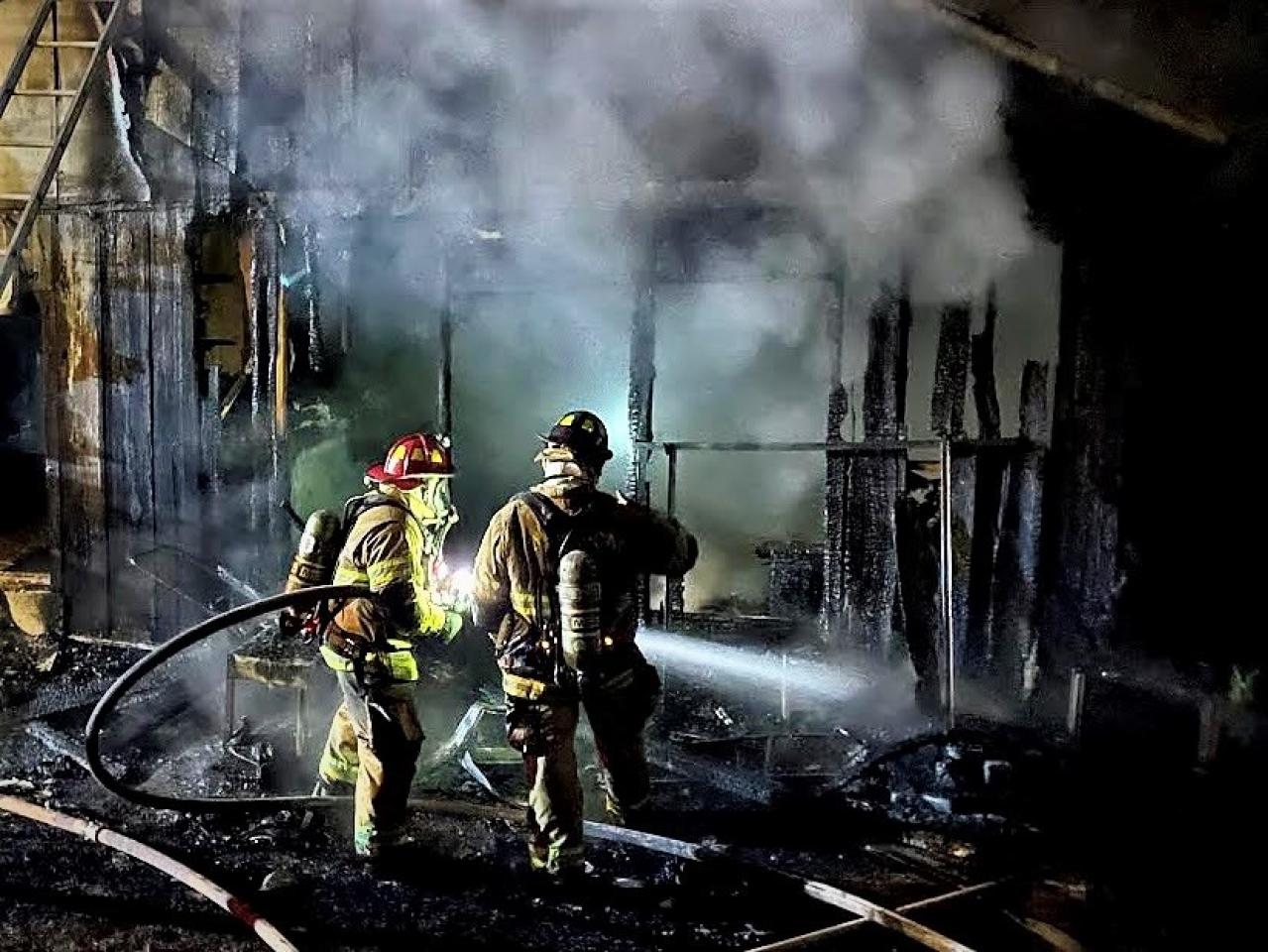 Dos bomberos con equipo completo extinguen un incendio nocturno en una estructura carbonizada y humeante, iluminados por las luces de sus cascos.