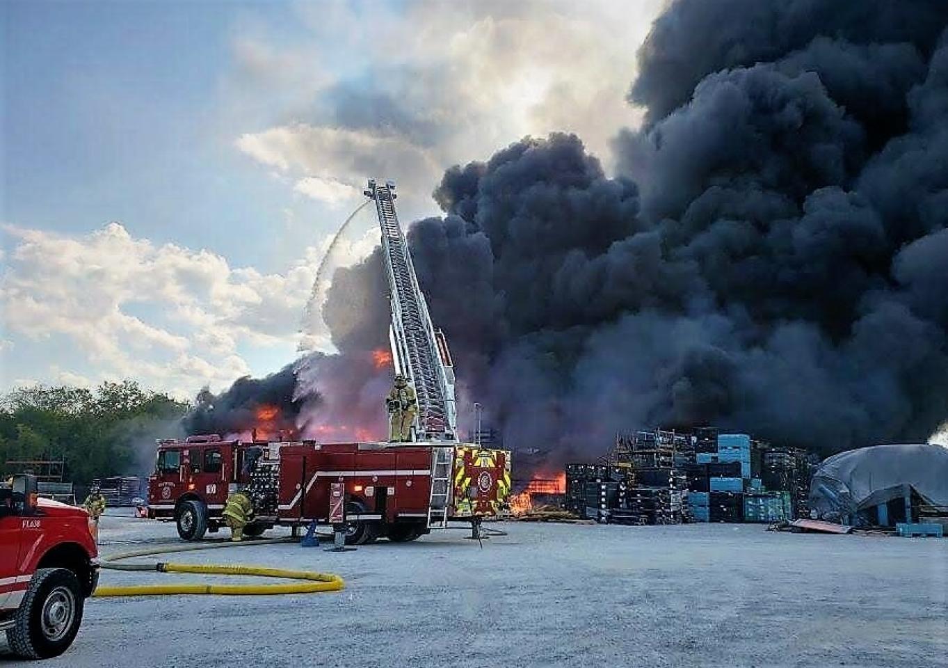 Bomberos combaten un gran incendio en un sitio industrial con humo negro espeso, utilizando un camión escalera y mangueras.