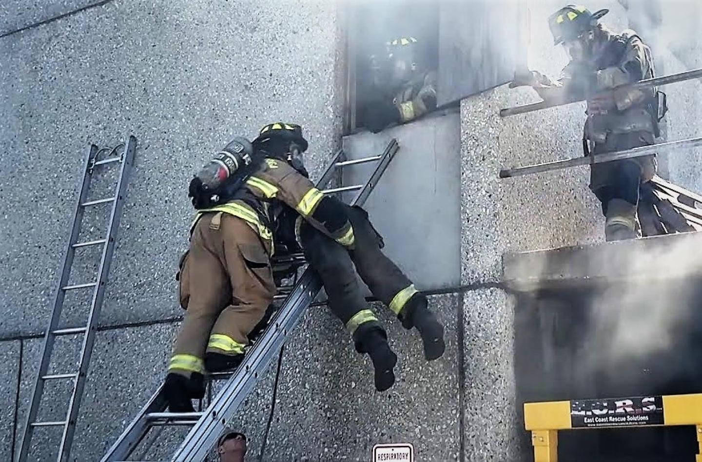 Firefighters in tan gear performing a rescue operation, carrying a person down a ladder from a smoke-filled building.