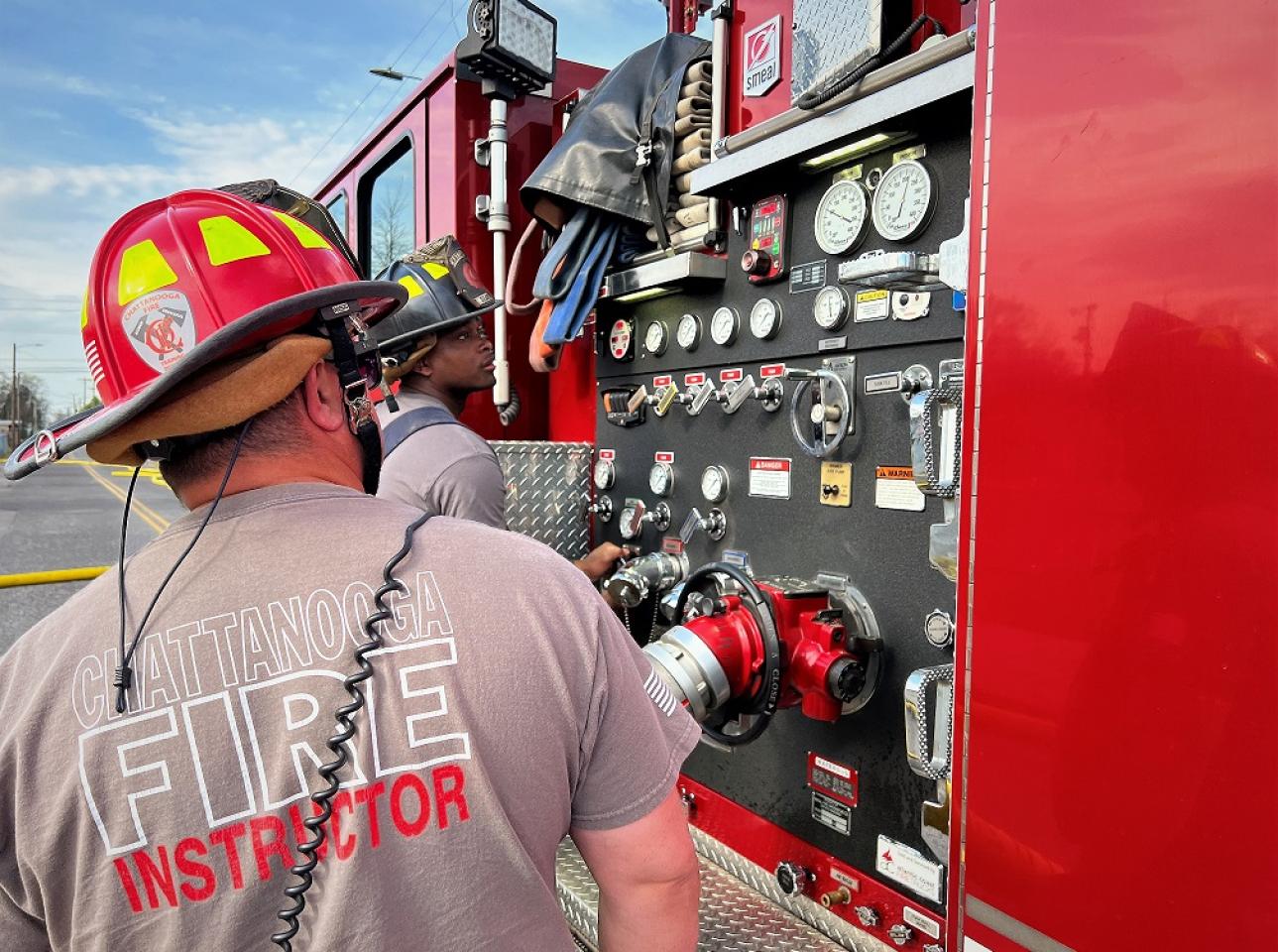 Fire instructor in a grey shirt and red helmet, guiding a firefighter in front of a fire truck's control panel during training