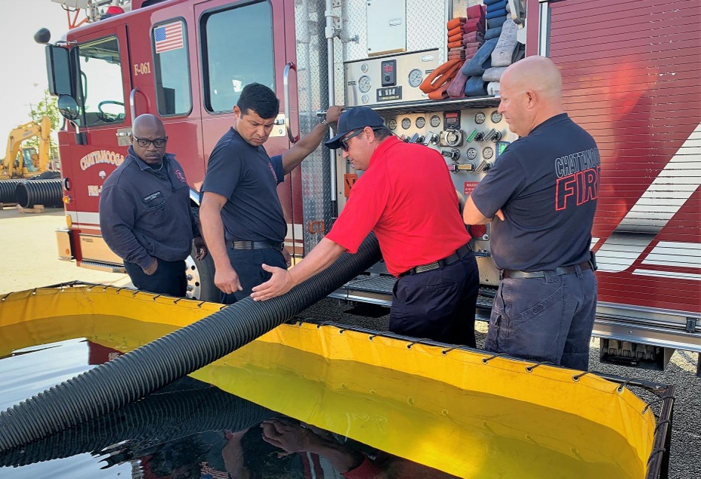 Firefighters in black and red shirts, working with a fire truck's equipment and hoses during a training exercise