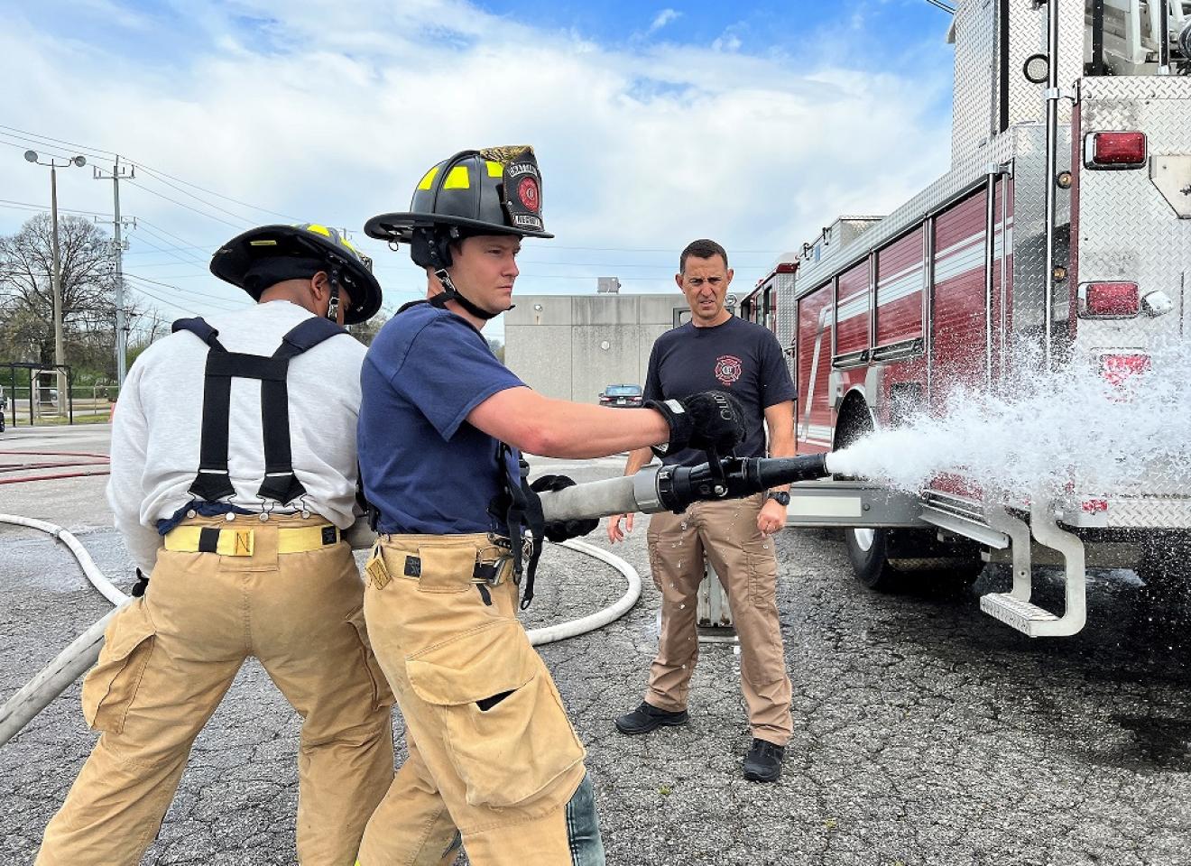 Two firefighters in training, with one holding a fire hose spraying water, supervised by an instructor in front of a fire truck