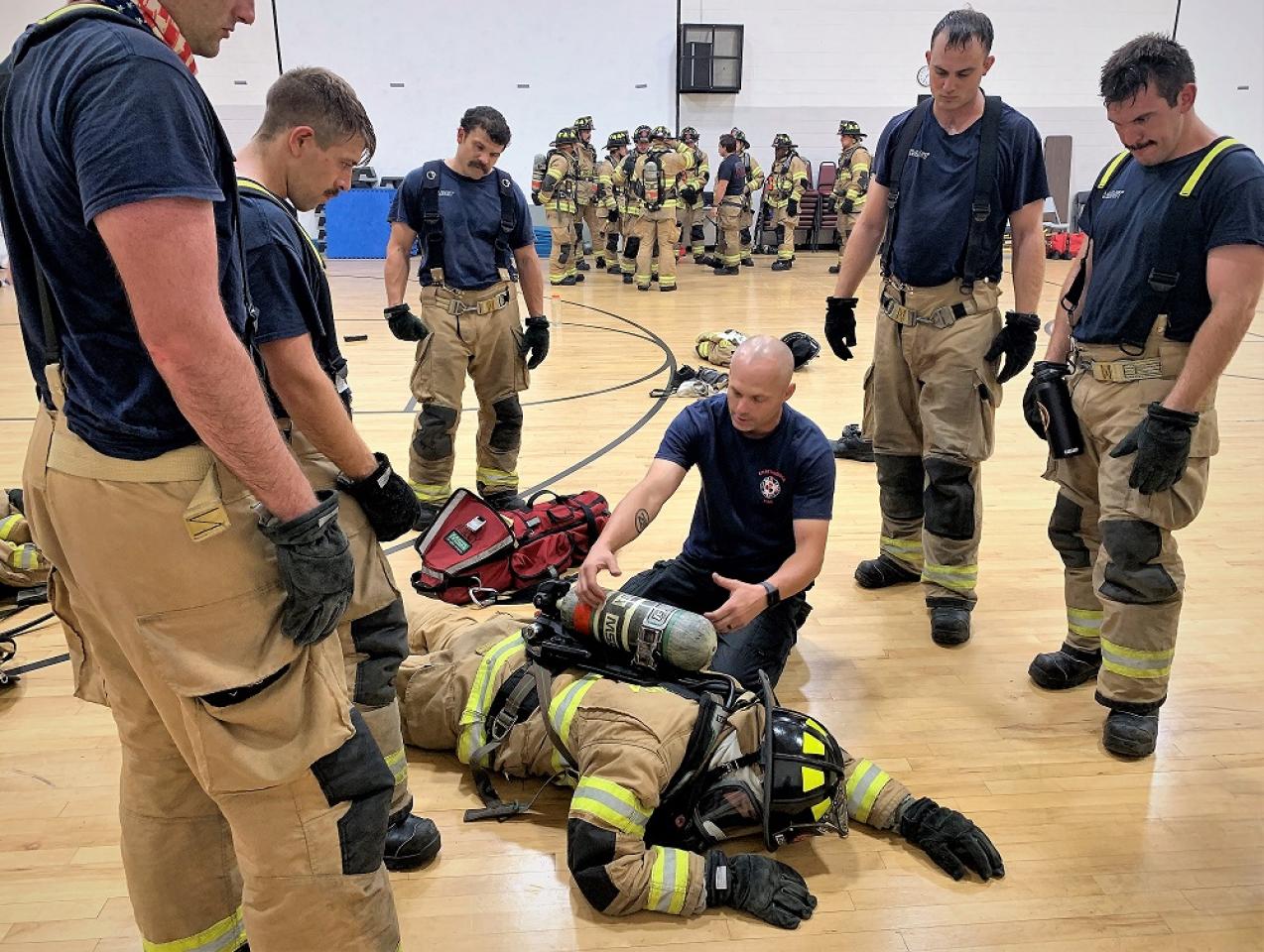 Group of firefighters in tan gear gathered around a firefighter lying on the floor in a gymnasium, receiving training