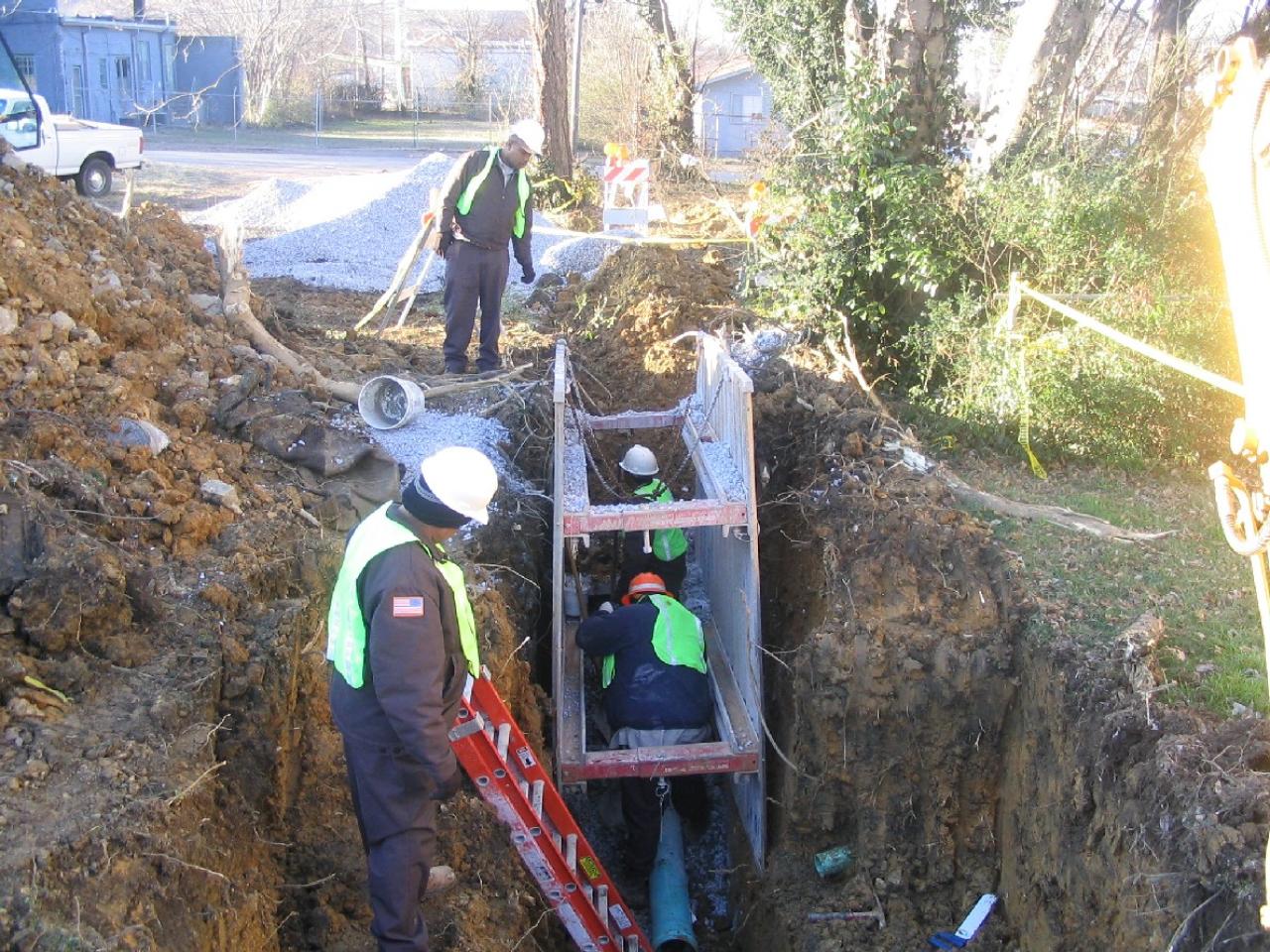 Three construction workers in green safety vests and hard hats working inside a metal trench box with surrounding dirt and an orange ladder