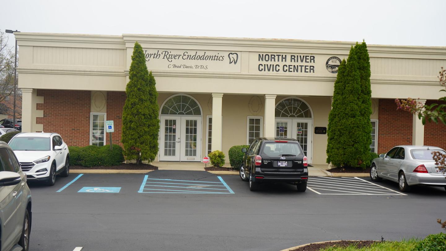 Front of North River Endodontics and Civic Center with beige facade, red brick sides, green bushes, and parked cars.