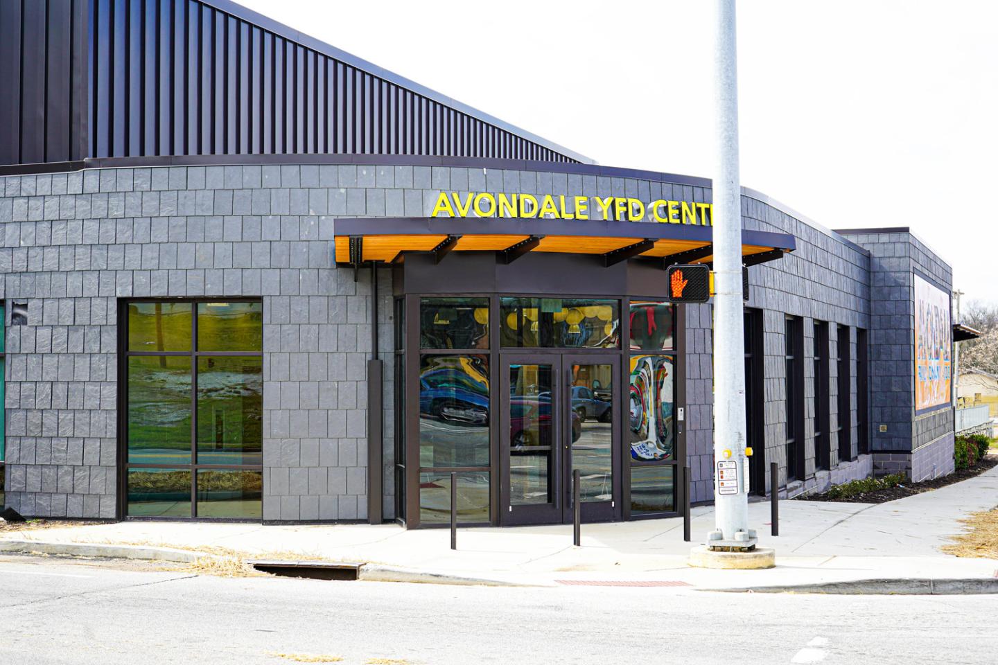 Fachada del Avondale Community Center con paredes de ladrillo gris, grandes puertas de vidrio y un letrero amarillo brillante bajo un dosel de madera.