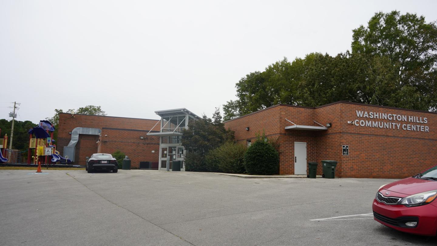 Washington Hills Community Center building with red brick exterior, a playground with colorful slides on the left, and a gray car parked in the lot. Trees with green foliage are in the background