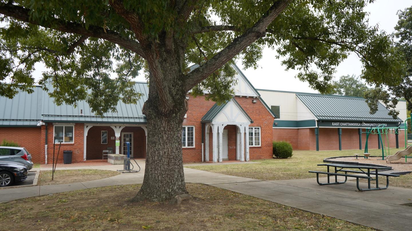 East Chattanooga Community Center with a large tree in the foreground. The brick building has a green metal roof and white accents, adjacent to a white building with a green roof. A playground and picnic table are visible on the right