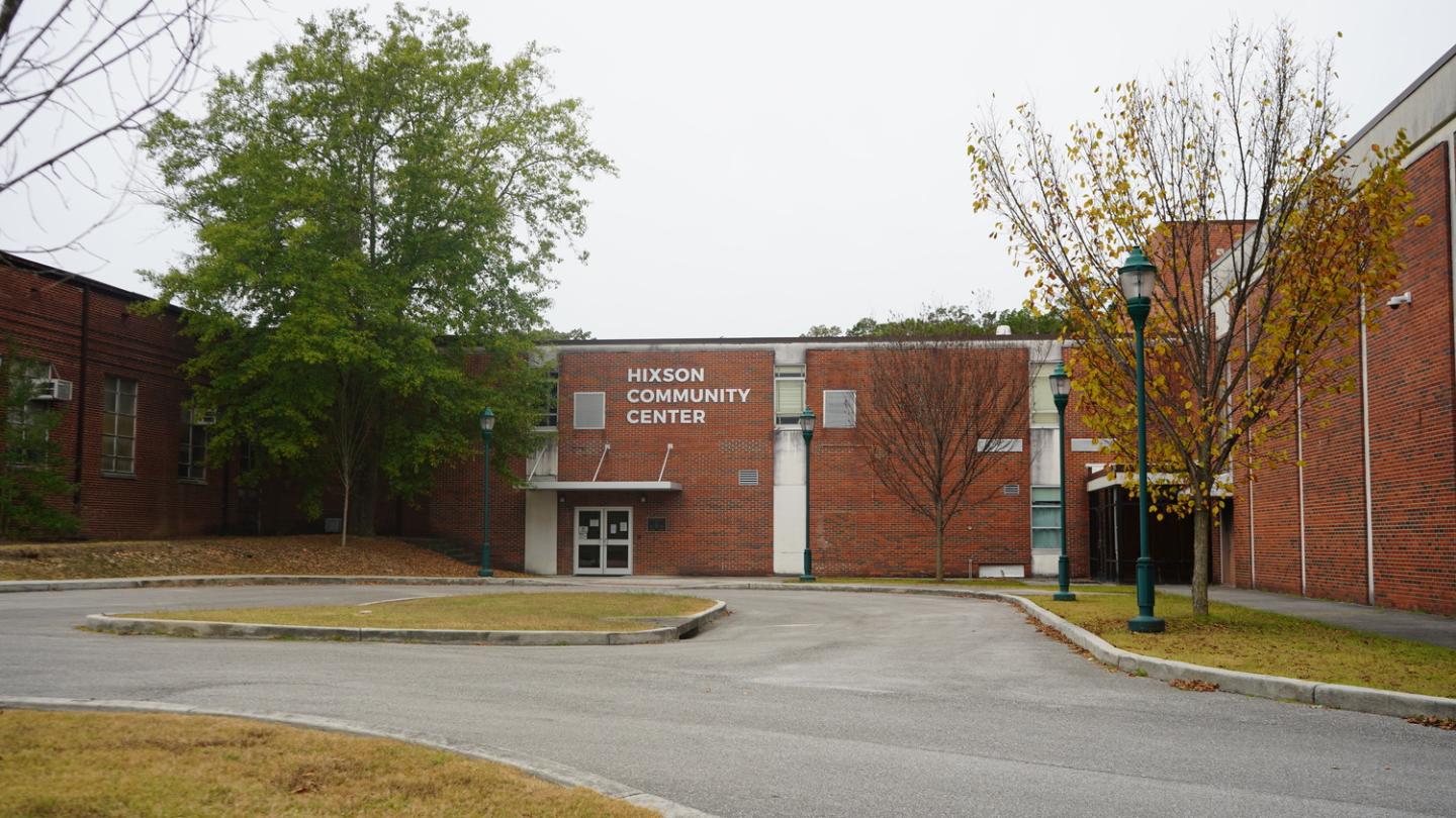 Front view of Hixson Community Center, a red brick building with a white sign on the facade. The scene features a gray, overcast sky with green and yellow autumn trees surrounding the center. A paved driveway curves towards the entrance