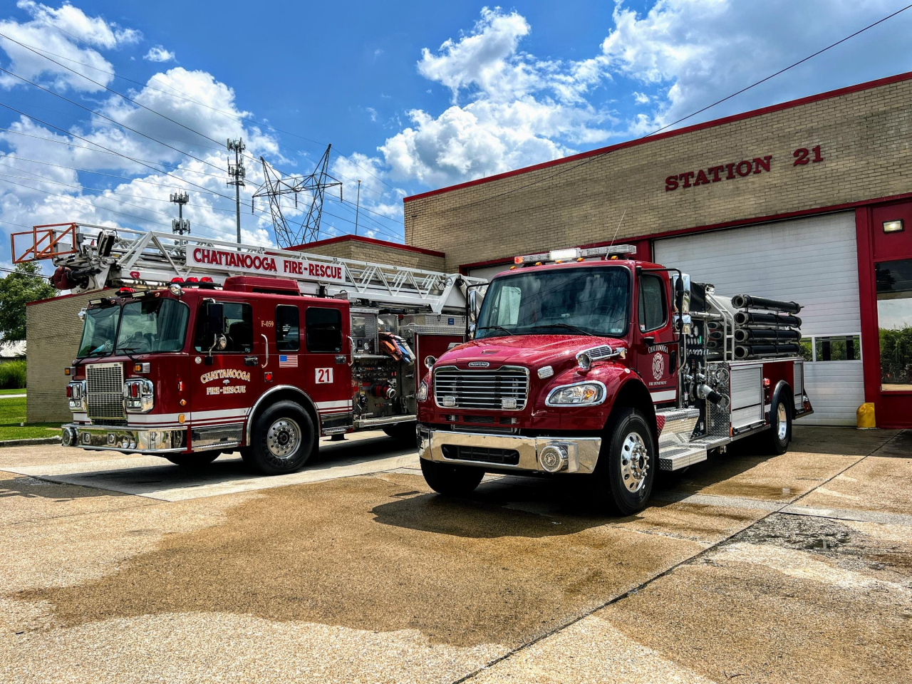 Exterior of Fire Station 21 with red brick walls, white garage doors, two fire trucks parked and a large fire department emblem on the front.