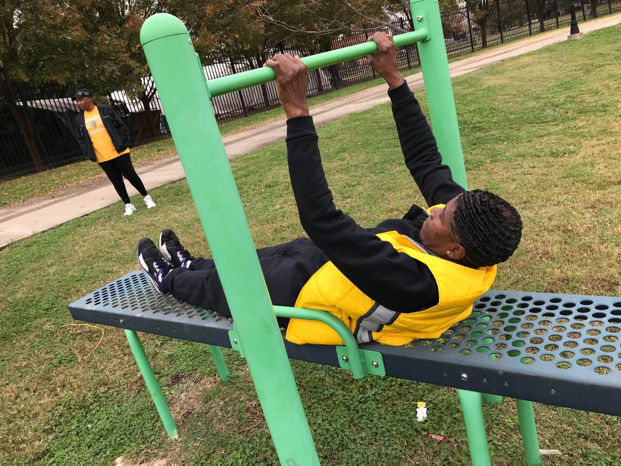 Two individuals exercising in an outdoor park. One person in the foreground is using green pull-up bars while lying on a perforated metal bench, wearing a yellow and gray jacket with black pants and sneakers. Another person in the background, dressed in a yellow shirt, black jacket, and black pants, stands near a pathway lined with grass and trees.