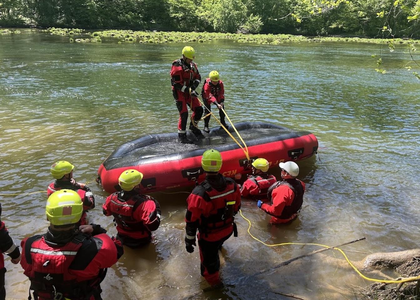Trabajadores de rescate con trajes rojos y negros y cascos amarillos realizan un ejercicio de entrenamiento en rescate acuático. Algunos están en una balsa inflable mientras otros están en el agua o en la orilla, sosteniendo cuerdas. El fondo incluye árboles y un río parcialmente iluminado por el sol.