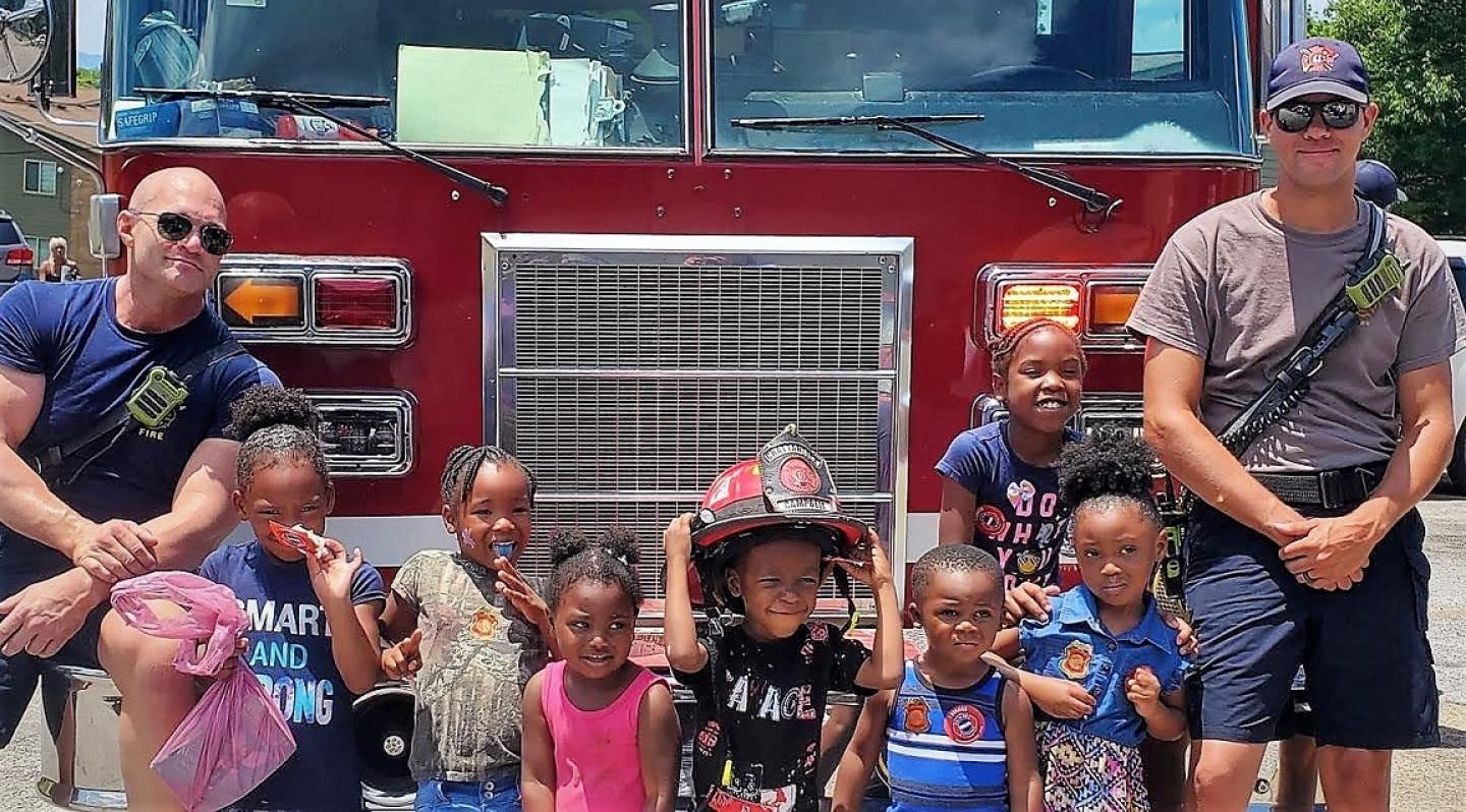 Dos bomberos con siete niños frente a un camión de bomberos sonriendo.
