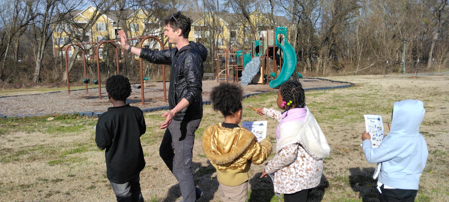 En esta imagen se observa a un grupo de niños en un entorno exterior, posiblemente un parque o patio escolar. Un adulto, de pie y con la mano extendida, parece estar dirigiendo la atención de los niños o explicando algo. Los niños, que varían en edad, están agrupados a su alrededor, algunos con tazas en las manos, y parecen estar participando en alguna actividad o lección al aire libre.