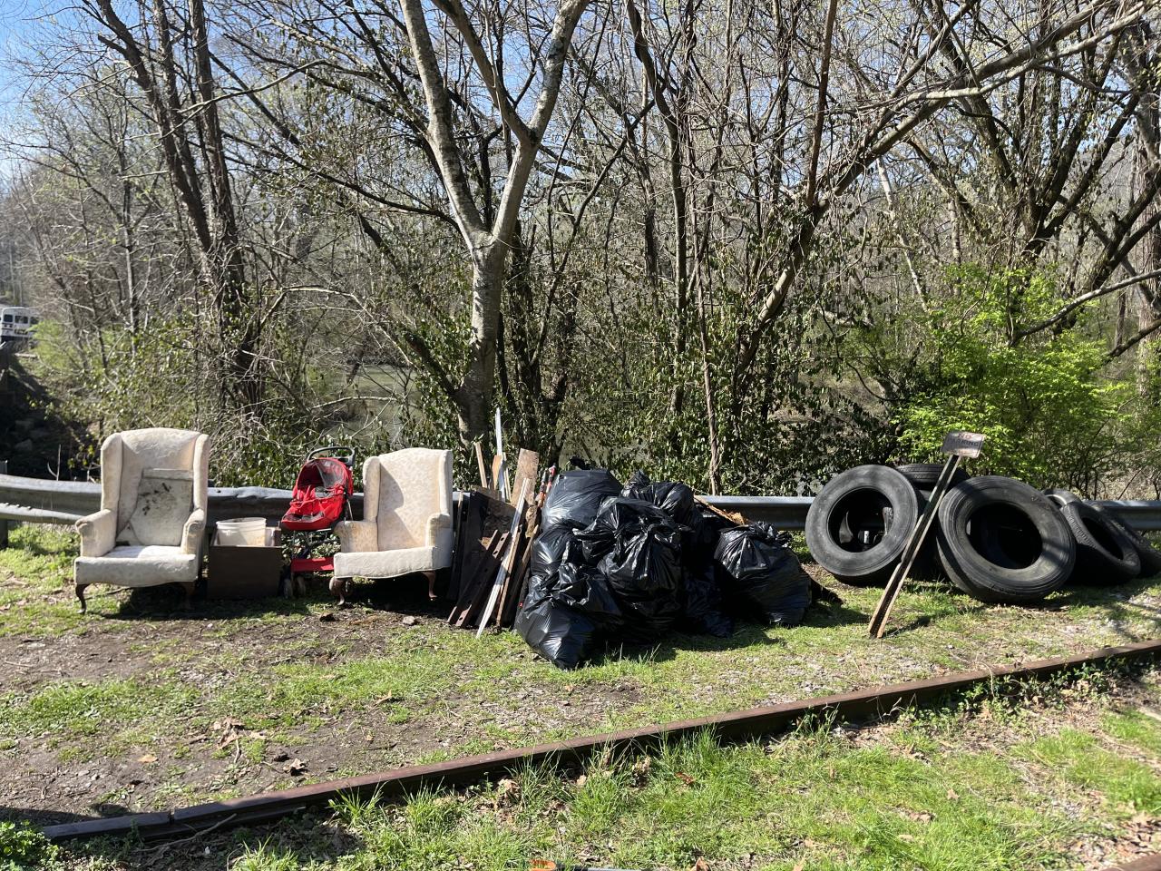 La imagen muestra una escena al aire libre con varios objetos apilados a lo largo de unas vías. En primer plano, se ven varios neumáticos apilados a la derecha. Hacia la izquierda, hay una bolsa de basura negra y lo que parecen ser algunos asientos o muebles.