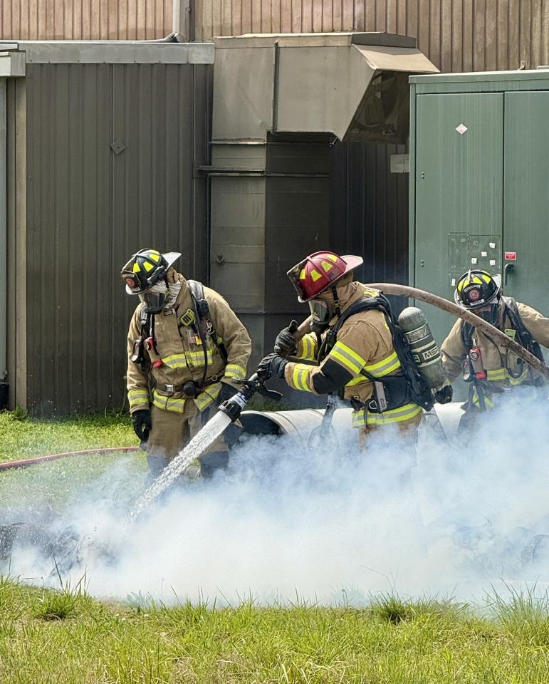 Three firefighters spray grass with water behind factory