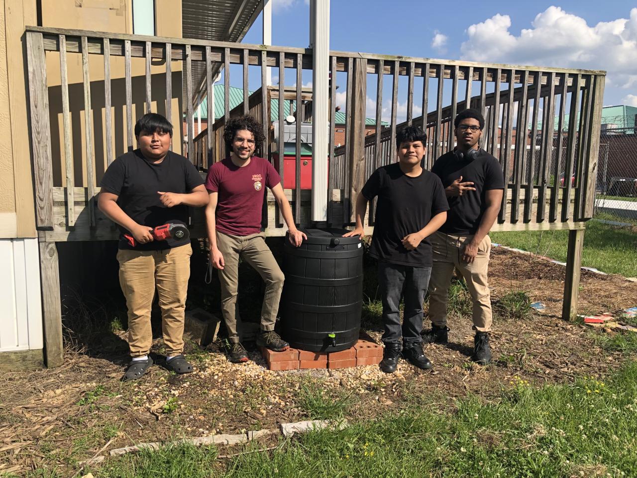 Un grupo de cuatro jóvenes está de pie junto a un gran barril negro, probablemente un recolector de agua de lluvia, frente a una estructura de madera tipo terraza.