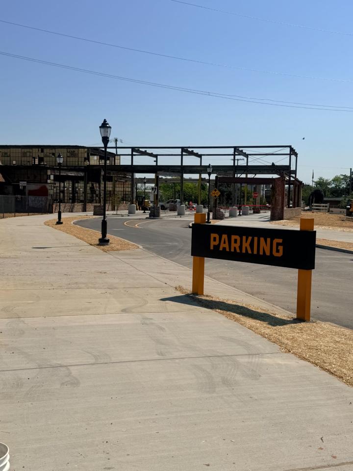 A 'Parking' sign stands next to a sidewalk leading toward an open-frame industrial building under construction with decorative black street lamps.