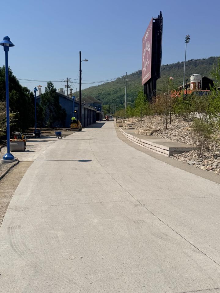 A wide concrete walkway at Erlanger Park, featuring tiered stone landscaping, a large maroon billboard, and a view of a green mountainside in the distance.