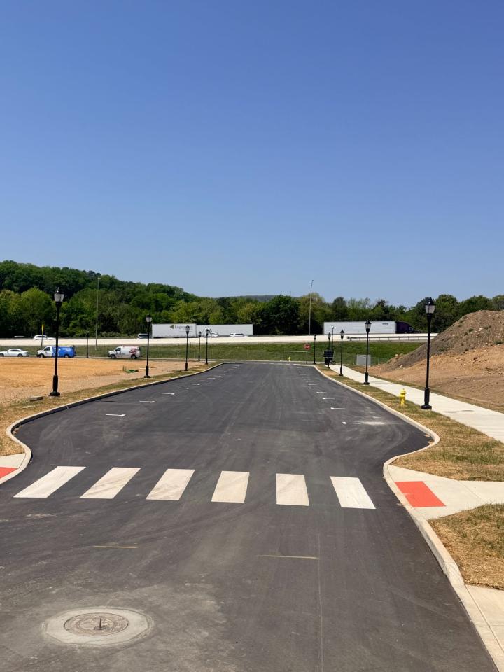 A newly paved road with a crosswalk and street lamps at a construction site, with a highway and green trees in the background
