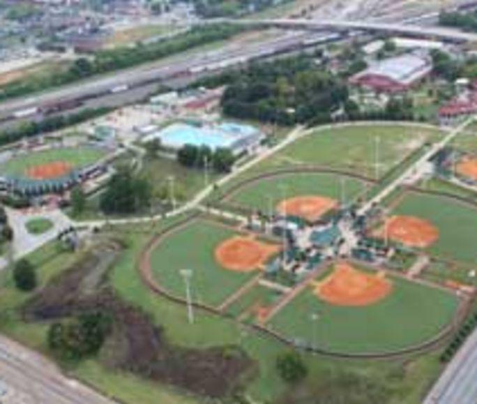 Aerial view of a large sports complex featuring multiple baseball fields with red infields and green outfields. The complex includes adjacent buildings, roads, and a swimming pool. Surrounding areas include various landscapes and infrastructure, including a highway. Aerial view of a large sports complex featuring multiple baseball fields with red infields and green outfields. The complex includes adjacent buildings, roads, and a swimming pool. Surrounding areas include various landscapes and infrastructure, including a highway.
