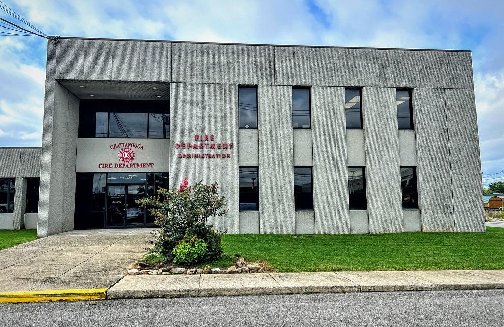 Edificio de concreto gris del Chattanooga Fire Department Administration con letrero rojo y un pequeño jardín al frente, que presenta césped verde y un arbusto con flores rosadas.