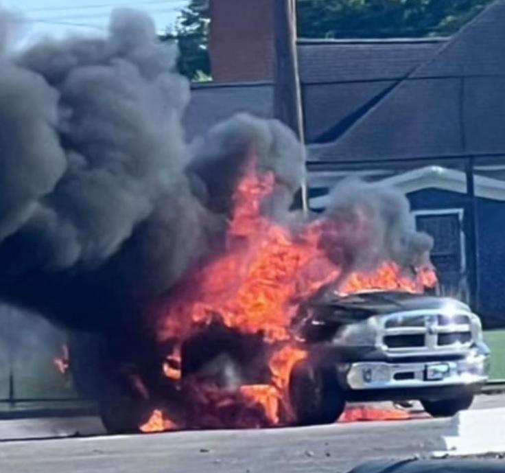 A truck is engulfed in flames on a street, emitting thick black smoke. The front end of the vehicle is completely ablaze. Nearby buildings and a power pole are visible in the background.