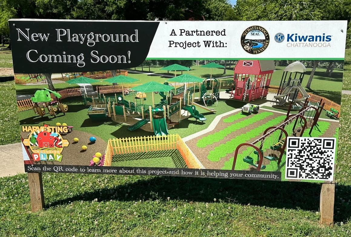 Un cartel en un parque anuncia un nuevo área de juegos. Presenta ilustraciones vívidas de equipos de juegos y múltiples logotipos, incluidos Harvesting Play, Kiwanis Chattanooga y otros. Se incluye un código QR para obtener más información sobre el proyecto.