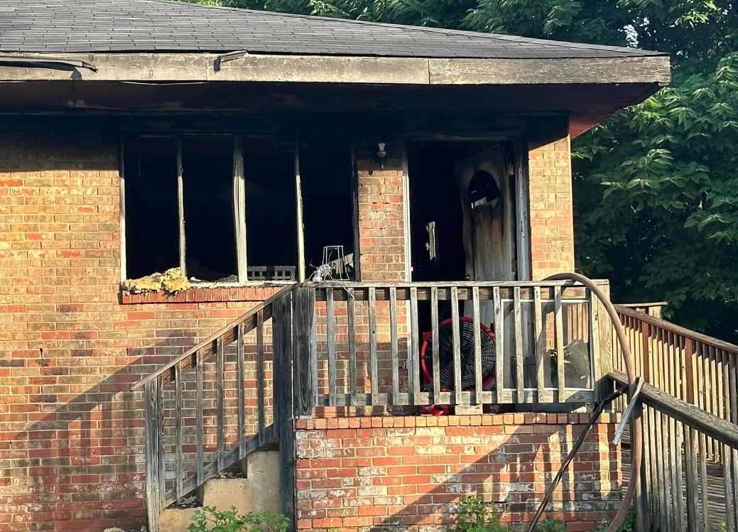 Imagen de una casa de ladrillo con daños significativos por incendio. Las ventanas están rotas y las paredes circundantes están chamuscadas. Los escalones y la barandilla de madera del frente permanecen intactos, pero muestran signos de desgaste. La puerta principal está parcialmente abierta, revelando más daños en el interior. Hay árboles en el fondo.