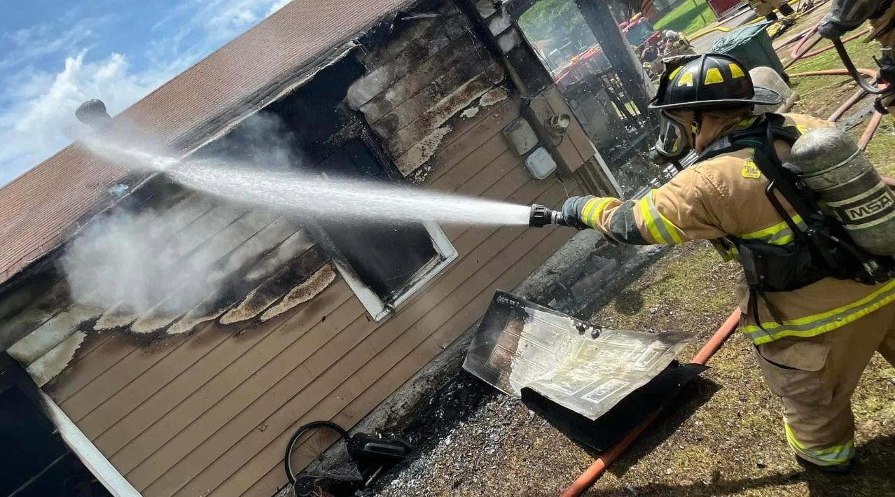 A firefighter in full gear sprays water from a hose onto a burning house with charred walls and smoke rising. The surrounding area is littered with debris, including a burnt piece of furniture. Other firefighters and equipment are visible in the background.