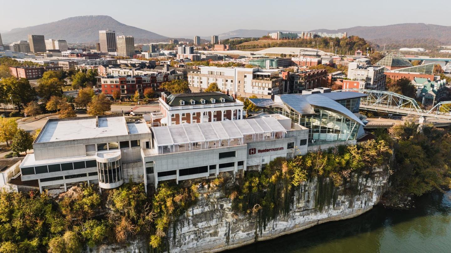 An aerial view of Chattanooga, showcasing the vibrant downtown area with modern buildings and green spaces. In the foreground, the Tennessee Aquarium is perched dramatically on a cliff overlooking the Tennessee River, with the iconic Walnut Street Bridge visible in the background.