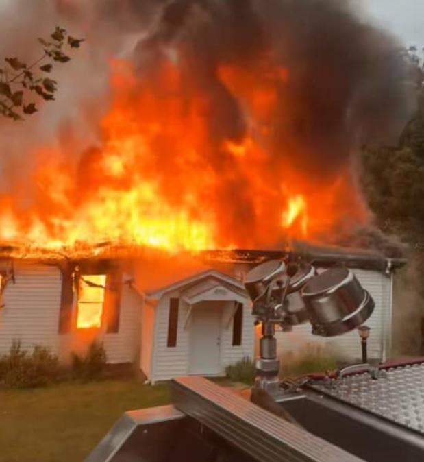 Una casa blanca envuelta en llamas con un denso humo negro elevándose hacia el cielo. El techo está casi completamente consumido por el fuego. Una parte de un camión de bomberos con luces montadas es visible en primer plano. Ramas de árboles se encuentran en la esquina superior izquierda.