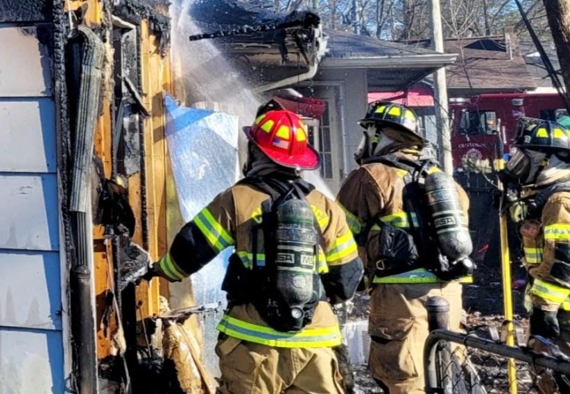 Three firefighters, wearing full gear and oxygen tanks, are extinguishing a house fire using hoses. The house's exterior shows heavy damage with charred walls. One firefighter with a red helmet directs water at the flames, while others assist. Fire truck visible in background.