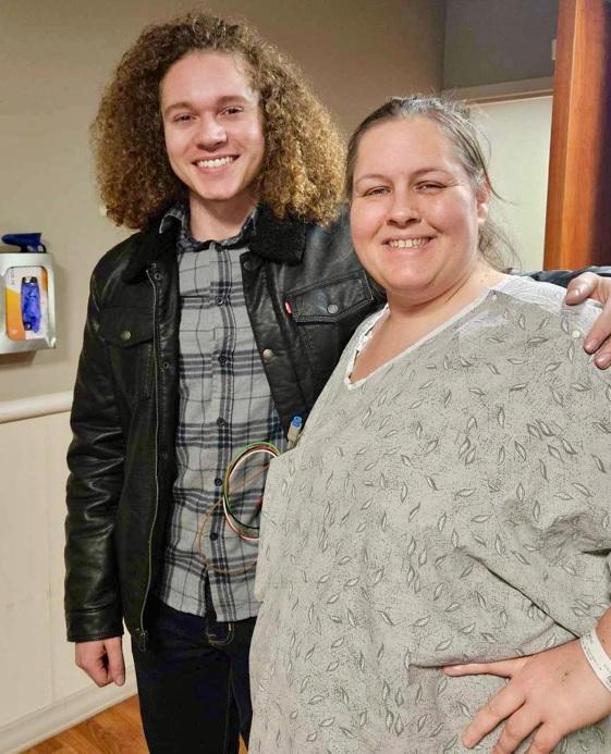 Un joven con cabello rizado, vistiendo una chaqueta de cuero, y una mujer con cabello largo, sonriendo y posando juntos en un entorno interior. La mujer lleva una blusa gris con estampado y tiene su brazo alrededor del hombro del hombre.