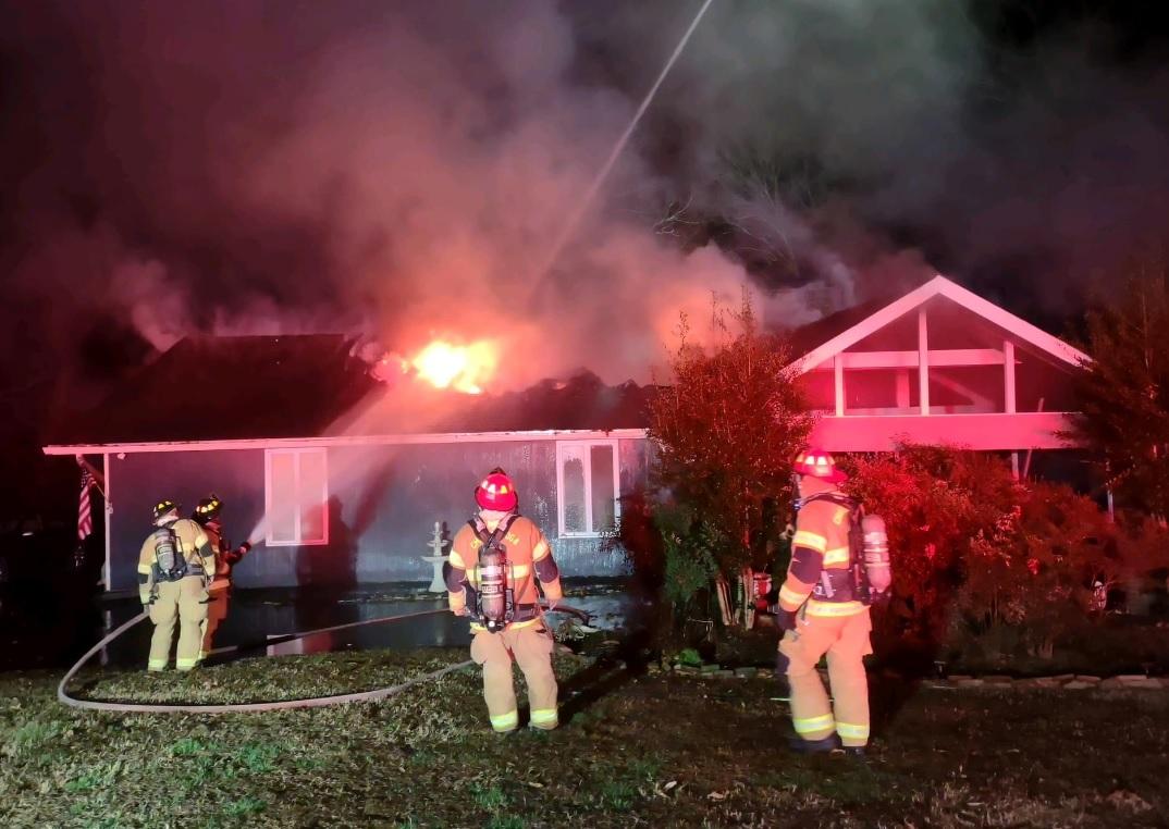 Bomberos con equipo de protección combaten un incendio en una casa durante la noche. Se ven llamas y humo saliendo del techo mientras un bombero utiliza una manguera. La escena está iluminada por las luces de los camiones de bomberos. La casa parece ser una residencia de un solo piso con daños significativos por el fuego.