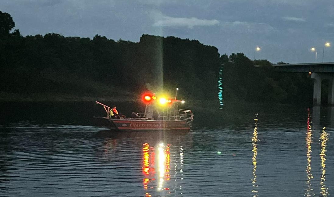 A boat with flashing red and yellow lights is on a calm body of water at night. The reflections of the lights shimmer on the water's surface. In the background, there are dark trees and a bridge with lights on under a dim sky.