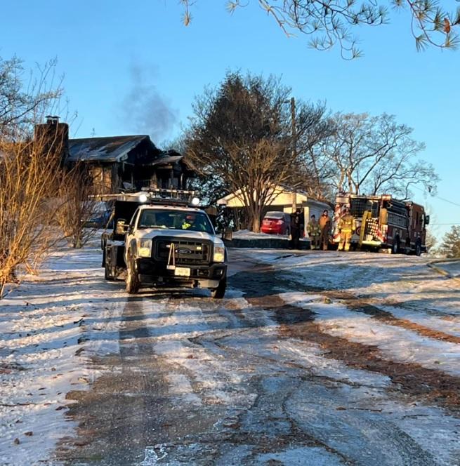 A fire truck and emergency responders are parked in front of a house with visible smoke damage on a snowy day. A second emergency vehicle is on the driveway leading up to the house. Snow-covered trees and bushes surround the property.