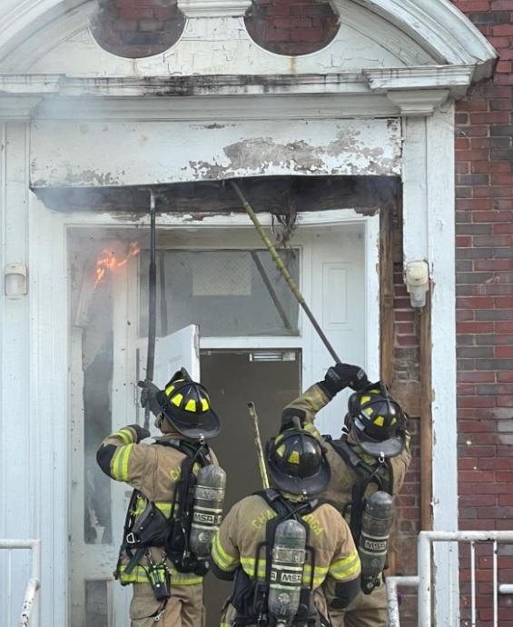 Three firefighters in full gear work to extinguish a fire near the entrance of a brick building. One firefighter sprays water while two others use tools to break apart the doorframe and remove burning material. Smoke and flames are visible above the door.