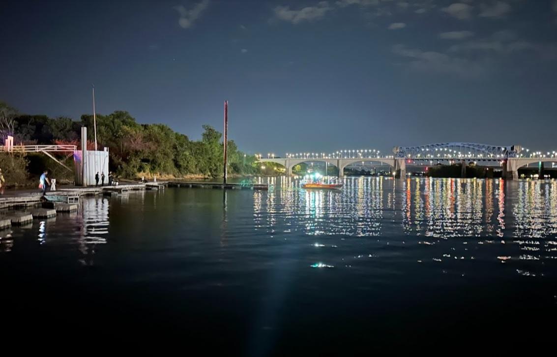 Nighttime view of a calm river with a brightly lit, arch-shaped bridge reflecting on the water's surface. There are docked boats and a few people at a pier on the left, with trees lining the riverbank. The sky is clear with some clouds.