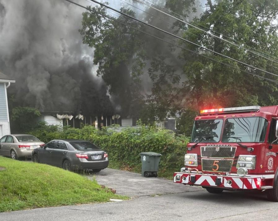 A residential house with thick black smoke billowing from it. Two cars are parked in the driveway, and a single green trash bin is visible. A fire truck, with red lights flashing and marked with the number 5, is parked on the street in front of the scene.