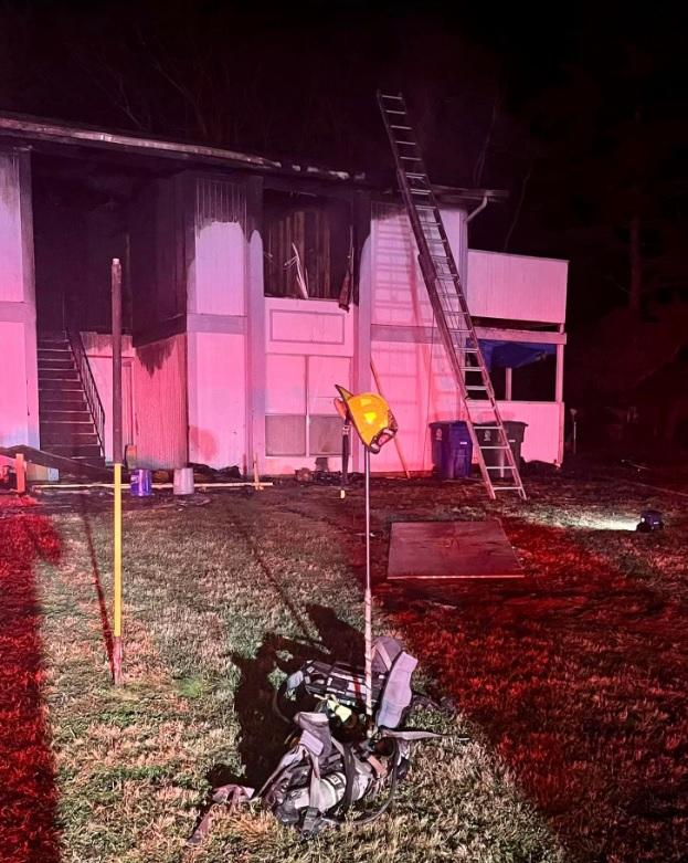 Un edificio de dos pisos se incendió, mostrando daños extensos en el piso superior. Una escalera está apoyada contra el edificio alcanzando una ventana del piso superior. Equipos de bomberos están esparcidos en el césped, iluminados por luces de emergencia rojas.