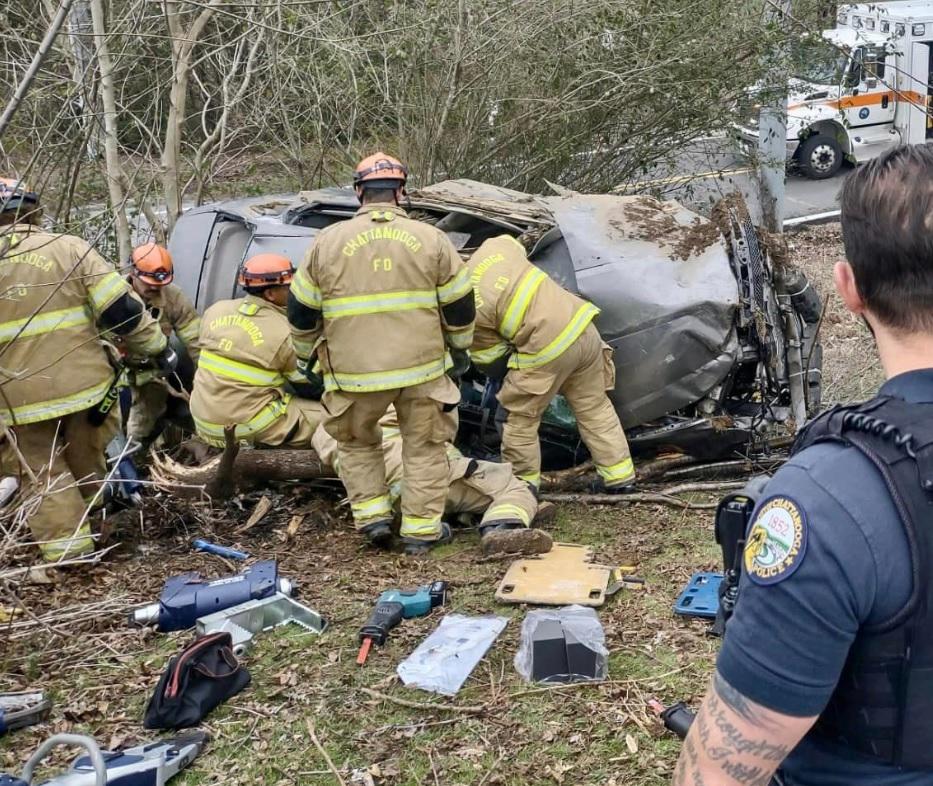 A group of firefighters in yellow suits and helmets are rescuing someone from a severely damaged car that has flipped over in a wooded area. A police officer stands in the foreground. Emergency equipment is scattered on the ground, and a fire truck is visible in the background.