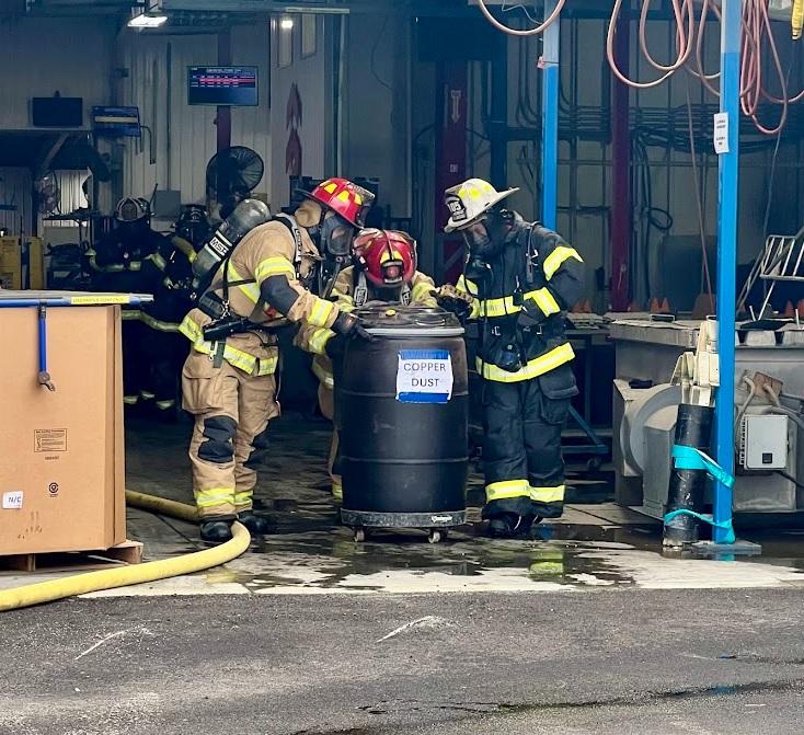 Tres bomberos con equipo completo y aparato de respiración inspeccionan un barril negro etiquetado como "Polvo de Cobre" dentro de un taller industrial. La escena incluye varios equipos y maquinaria, una caja grande a la izquierda y mangueras enrolladas colgando del techo.