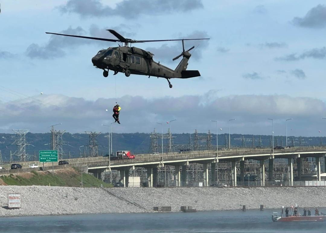 Un helicóptero se mantiene en el aire sobre un cuerpo de agua con una persona suspendida debajo, probablemente durante una operación de rescate. En el fondo, hay una zona industrial con líneas eléctricas y un puente que cruza el agua. Se ve un barco cerca de la orilla del agua.